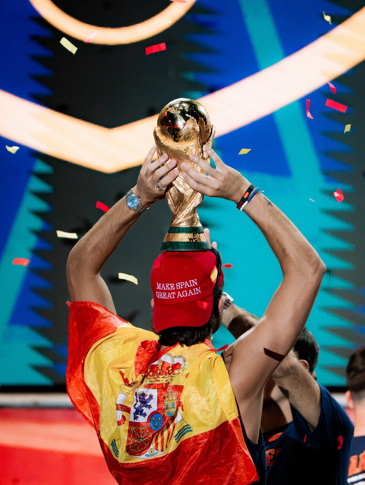 Ferran Torres in a red and yellow Spain jersey and red make spain great again hat holds a large trophy above their head on a stage with confetti and a blue geometric backdrop.
